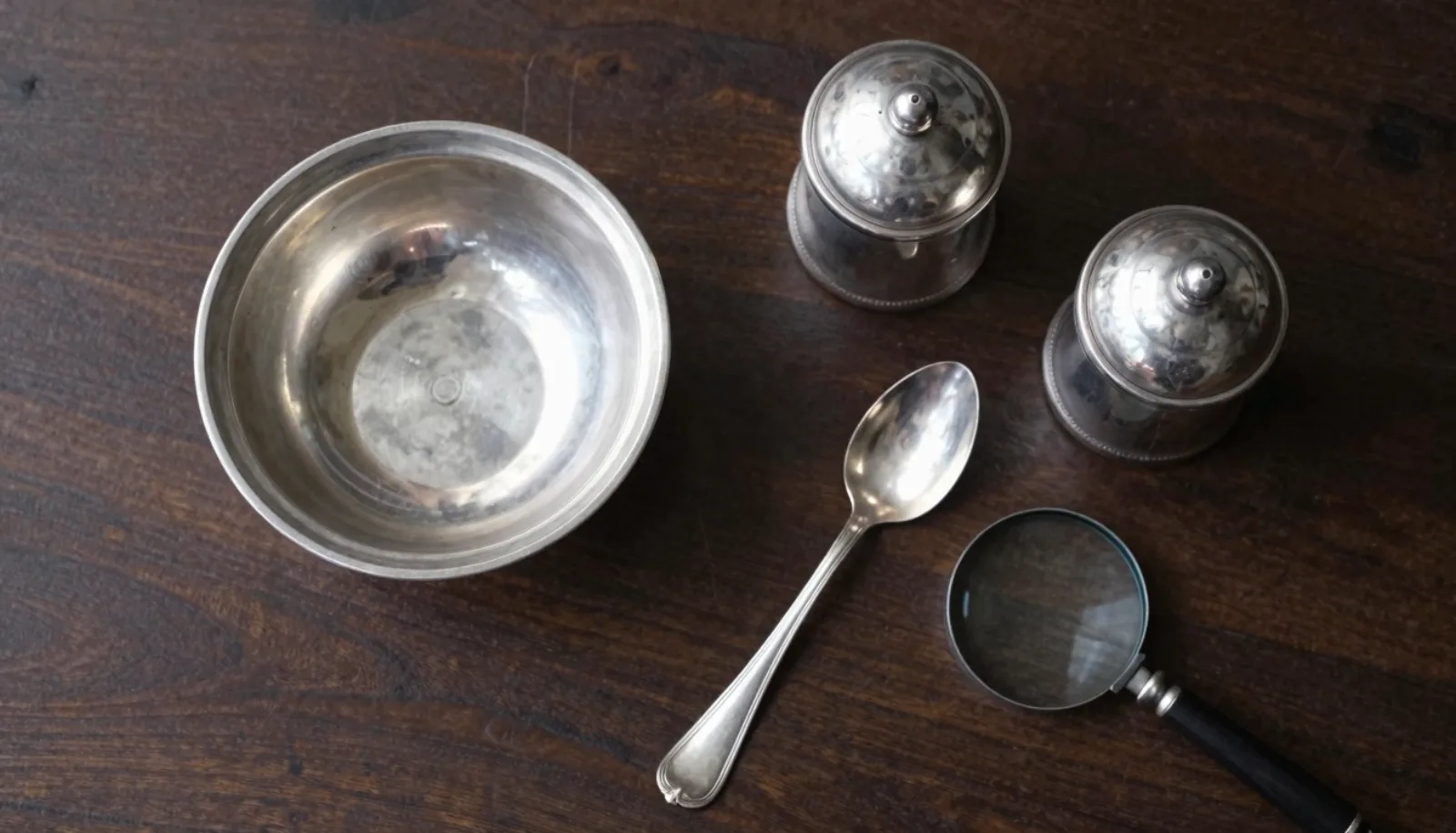 Antique sterling silver bowl, pair of salt cellars and a spoon on dark wood with a jeweller’s loupe